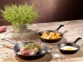 Trois poêles en fonte de diamètres différents posées sur une table en bois à côté d'herbes aromatiques, chacune contenant un plat cuisiné différent : oeuf au plat, filet mignon et patates sautées
