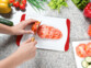 Mise en situation d'une personne découpant une tomate coeur de boeuf sur une planche à découper blanche et rouge sur un plan de travail en granit à côté de légumes