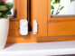 Mini alarme VisorTech mise en place sur le rebord extérieur d'une fenêtre en bois, à côté d'un pot de fleur blanc cachant le dispositif anti-intrusion, fixé par bande adhésive ou vis et alimenté par piles bouton LR44