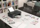 Mise en situation de l'imprimante jet d'encre Canon Pixma TS3550i posée sur une table de travail dans une salle d'étude avec une multitude de documents éparpillés sur la table