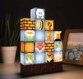 Lampe de décoration interactive et personnalisable Super Mario Bros Build a Level posée dans l'obscurité d'une pièce sur une table à côté d'une plante en pot et d'un cadre photo mettant en scène un port