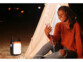 Jeune femme noire souriante portant un pull marron et un jean troué au niveau des genoux assise devant une tente éclairée par la batterie externe à dynamo sur le sable et regardant son téléphone mobile branché à cette même batterie de secours