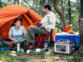 Homme et femme cinquantenaires en camping dans une forêt, devant une tente orange avec leur glacière bleu et blanche alimentée par la batterie nomade HSG-1200