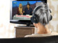 Femme âgée les cheveux gris attachées et posée sur un canapé noir de dos regardant les informations à la télévision avec le casque et amplificateur de son sans fil OK-330.hs