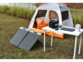 Panneau solaire pliable déplié au sol avec tente de camping derrière et table avec batterie nomade posée à côté, le tout le long d'un canal