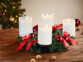 Couronne de l'Avent coloris rouge avec 4 bougies LED blanches posée sur une table de salon en bois à côté de noix entières devant un sapin de Noël décoré de guirlandes lumineuses et décorations de Noël