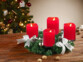 Couronne de l'Avent coloris argent avec 4 bougies factices LED rouges posée sur une table de salon en bois à côté de noix entières devant un sapin de Noël décoré de guirlandes lumineuses et décorations de Noël