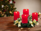 Couronne de l'Avent coloris doré avec 4 bougies LED coloris rouge posée sur une table de salon en bois à côté de noix entières devant un sapin de Noël décoré de guirlandes lumineuses et décorations de Noël