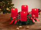 Couronne de l'Avent coloris rouge avec 4 fausses bougies LED rouges posée sur une table de salon en bois à côté de noix entières devant un sapin de Noël décoré de guirlandes lumineuses et décorations de Noël