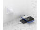 Mise en situation de la batterie externe noire posée au sol, branchée à une prise murale par le biais d'un adaptateur secteur et d'un câble USB blancs et rechargeant simultanément un smartphone sans fil