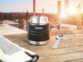 Mise en situation d'un mug isotherme ouvert posé sur une table en bois à côté de sa cuillère en inox avec un coucher de soleil en arrière-plan