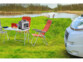 Mise en situation du générateur solaire HSG-1220 posé sur une table de camping entourée de deux chaises rouges devant un camping-car garé sur l'herbe devant un plan d'eau pour pêcher