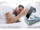 Chargement en cours d'une Apple Watch, d'un iPhone et du boîtier d'une paire d'EarPods sur la station de chargement posé sur un livre sur une table de nuit à côté d'un homme dormant dans un lit aux draps blancs